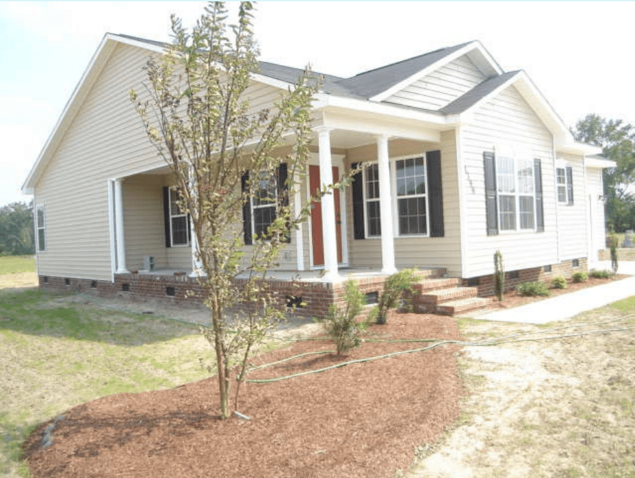 A new single-family home with light beige siding, white columns, and black shutters, featuring a landscaped front yard with a small tree and surrounding mulch.