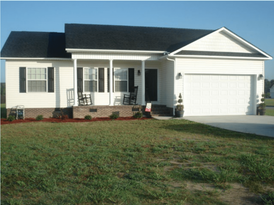 Front view of a single-story house with a black roof, white siding, and a brick foundation. The house features a porch with rocking chairs, two windows, and a garage.