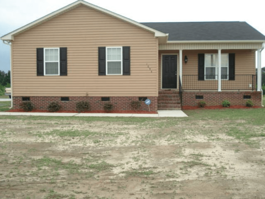 Single-story home with beige vinyl siding, black shutters, and a front porch featuring brick steps and landscaping.