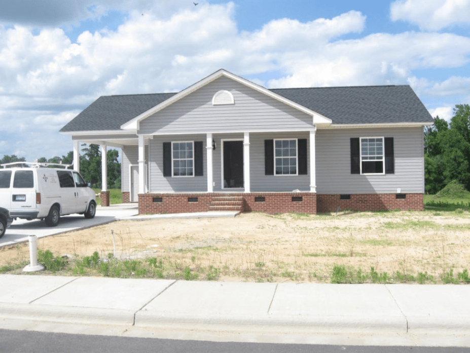 Front view of a newly constructed single-family home with gray siding, black shutters, and a brick foundation, located on a vacant lot.