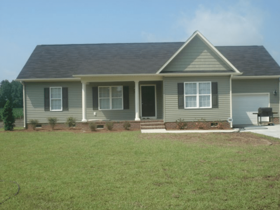 A single-story house with green siding, a black roof, and white trim, featuring a front porch with columns, two windows on the left, and a garage on the right.