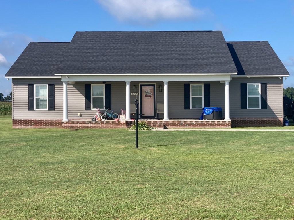 Front view of a single-story home with a black roof, beige siding, and a brick foundation, featuring a porch and green lawn.