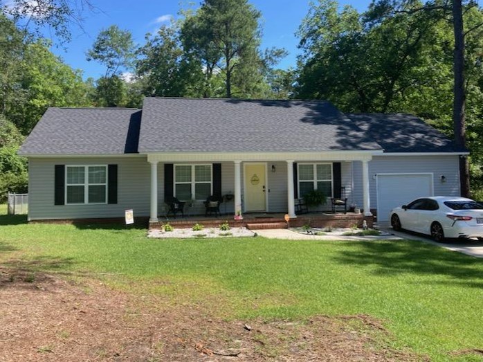 A single-story house with a gray exterior, black shutters, and a well-kept lawn. The front porch features two chairs and is adorned with flower beds, while a white car is parked in the driveway.